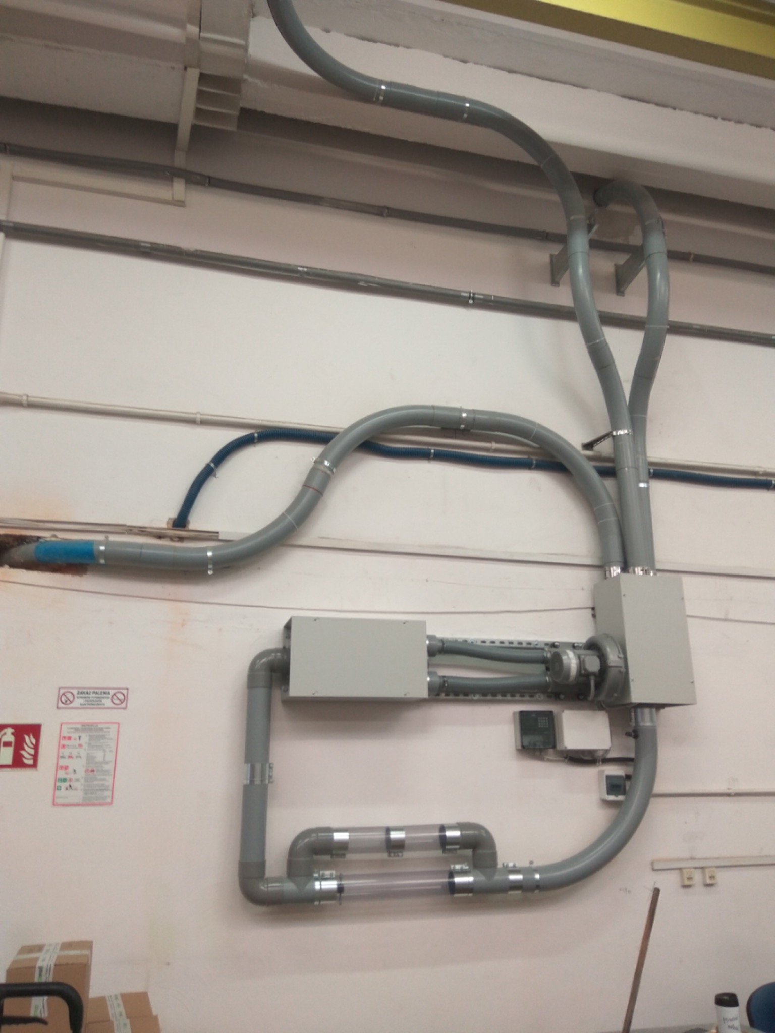 Szare rury wentylacyjne z elementami przezroczystymi, zamontowane na ścianie w hali przemysłowej, z widocznymi puszkami przyłączeniowymi i fragmentami instalacji elektrycznej.