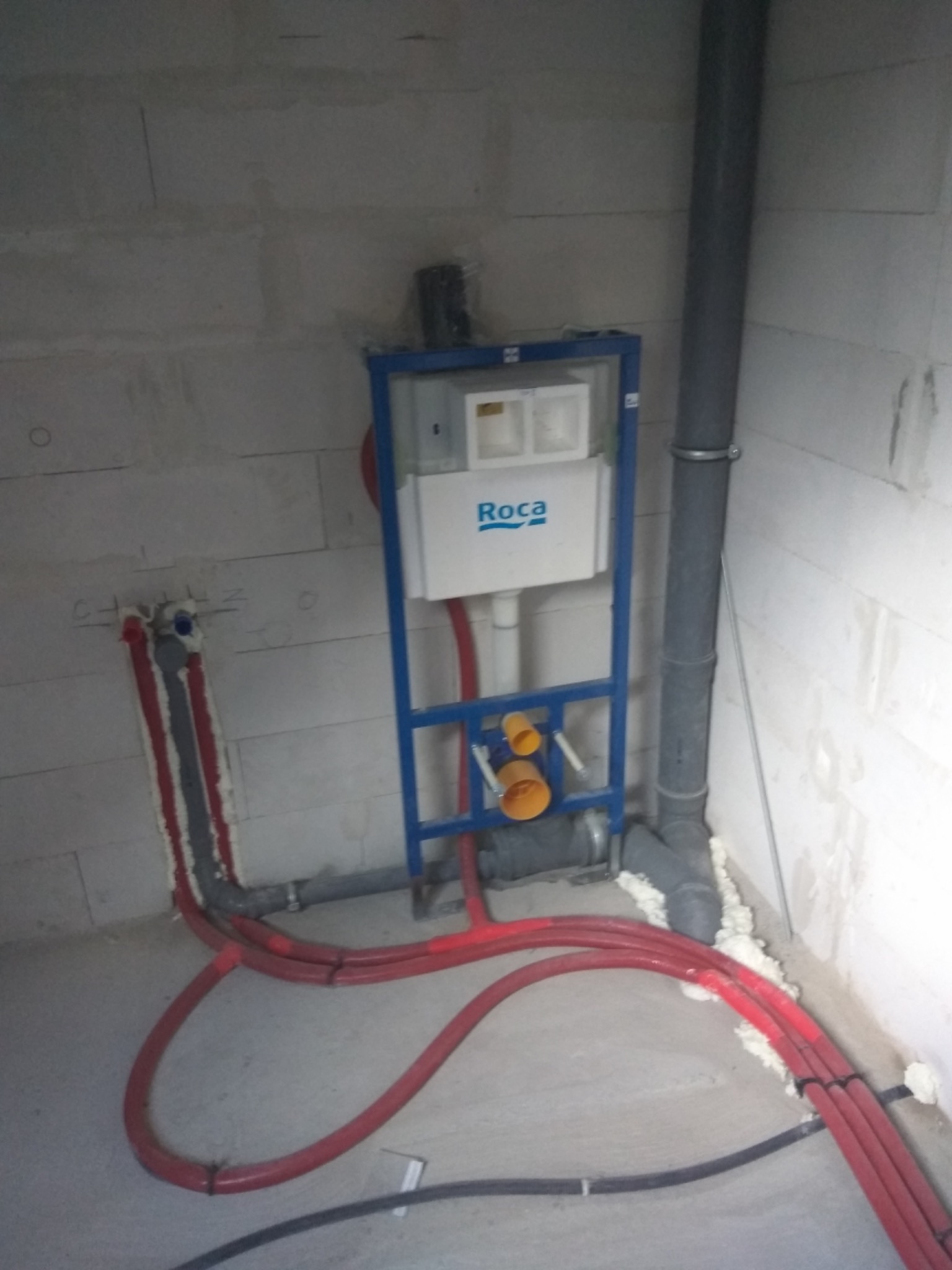 Stelaż podtynkowy WC marki Roca zamontowany na tle surowych ścian z betonu komórkowego, widoczne czerwone rury instalacji wodnej i szara rura kanalizacyjna, całość zabezpieczona pianką montażową.