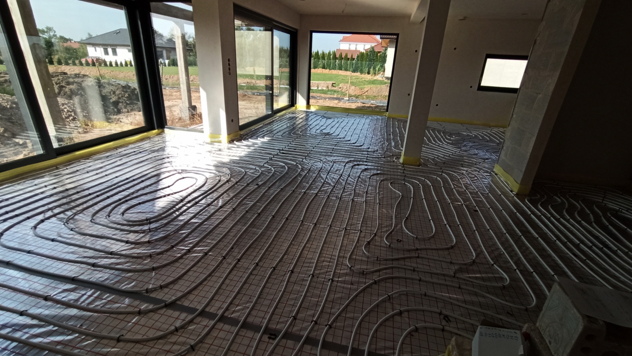 Rozłożona instalacja ogrzewania podłogowego na siatce w surowym wnętrzu domu z dużymi oknami tarasowymi i widokiem na ogród.