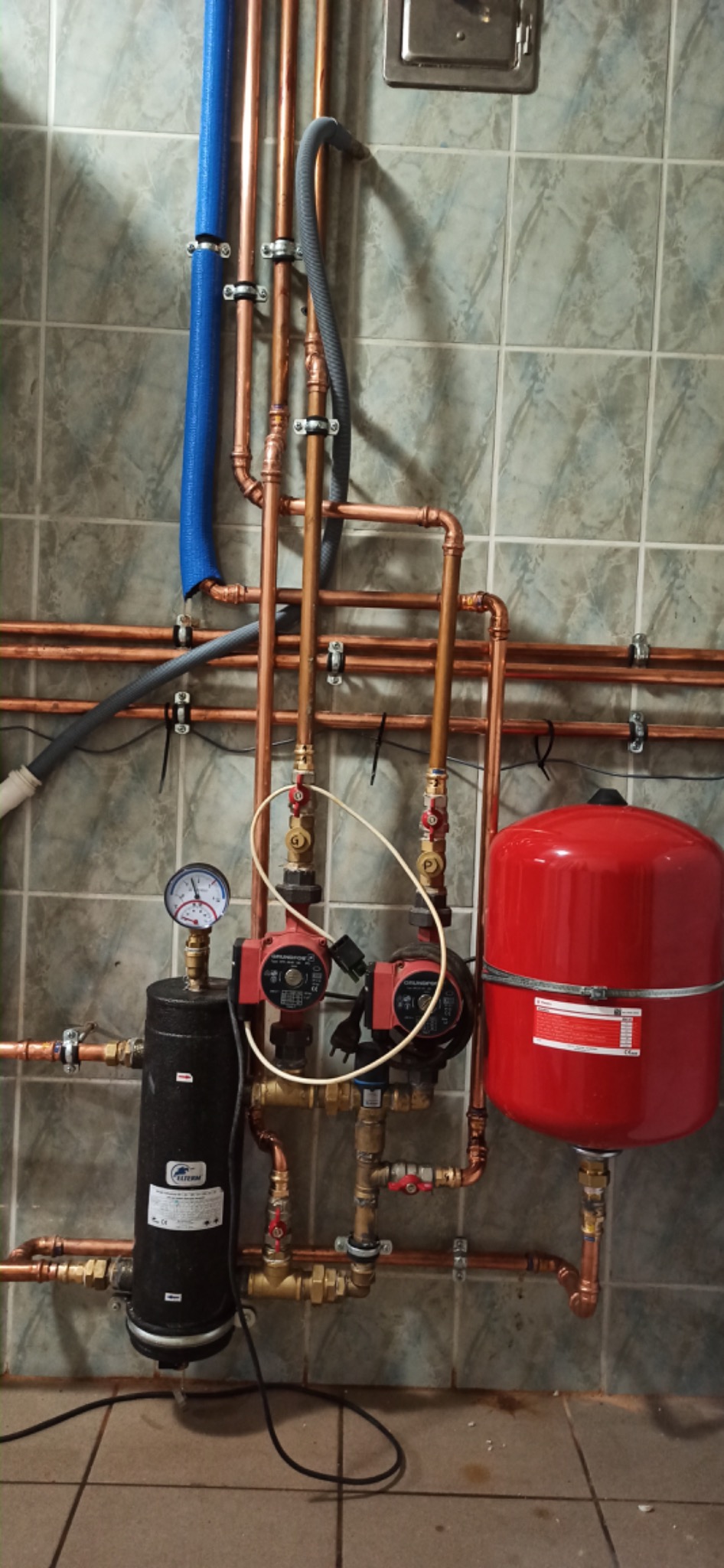 Nowa instalacja centralnego ogrzewania z miedzianych rur, pomp obiegowych, czarnego filtra i czerwonego zbiornika wyrównawczego zamontowana na ścianie z szarych płytek.