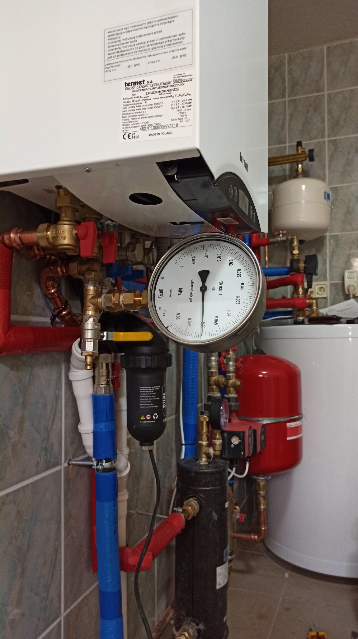 Kocioł gazowy kondensacyjny marki Termet z zainstalowanym manometrem, widoczne rury instalacji CO w kolorze czerwonym i niebieskim, filtr wody, oraz elementy instalacji grzewczej.