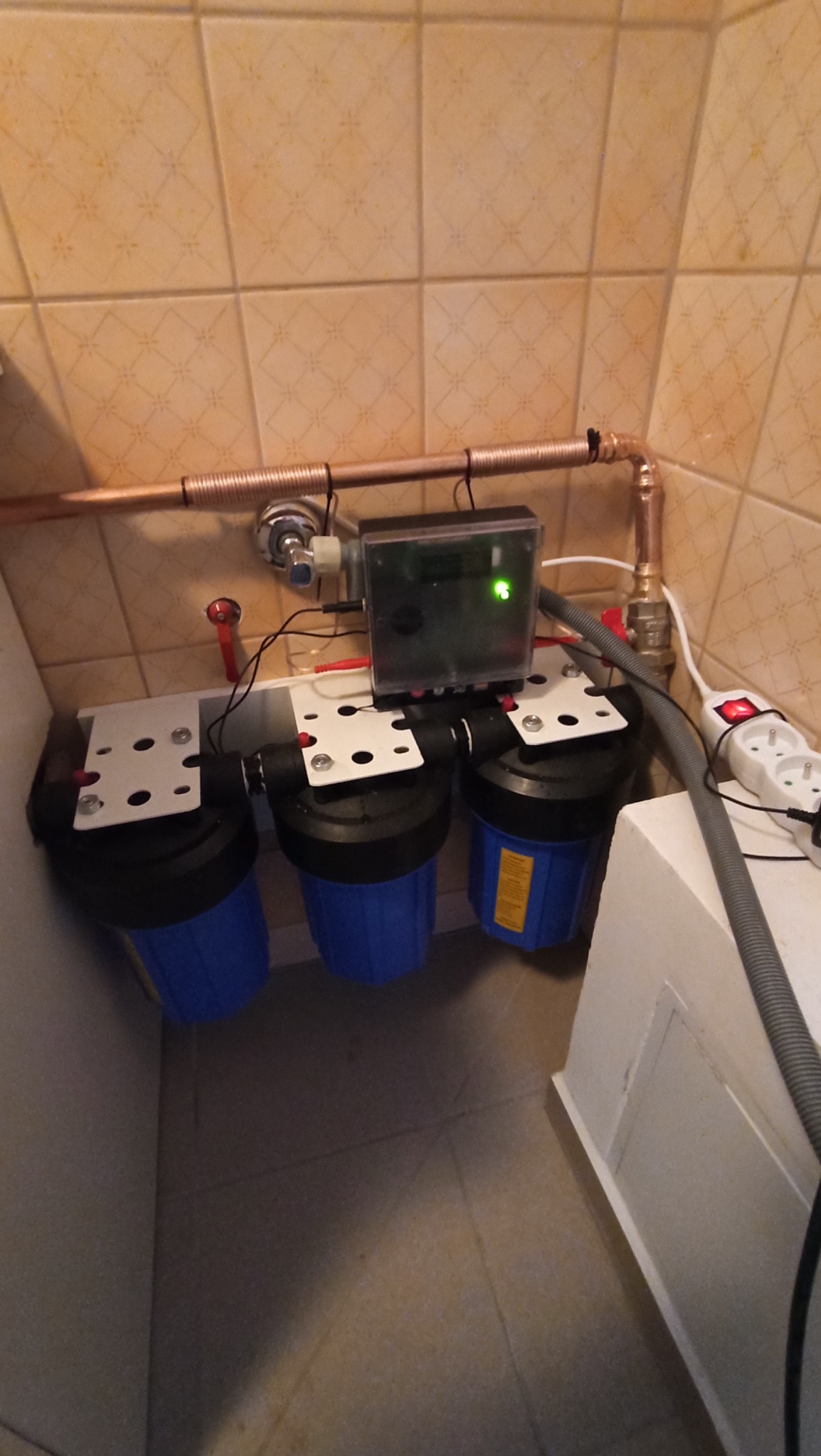 Trzy niebieskie filtry wody podłączone do miedzianej rury z zaworem, zamontowane w pomieszczeniu z beżowymi płytkami, widoczny panel sterujący i gniazdko elektryczne z włączonym przedłużaczem.