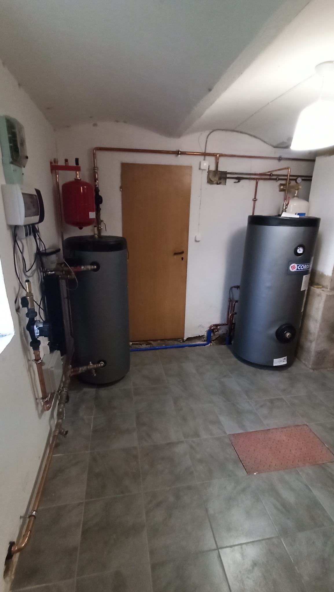 Dwa szare zasobniki ciepłej wody użytkowej w kotłowni, z widocznymi rurami miedzianymi i elementami instalacji. Na podłodze szare płytki imitujące kamień.