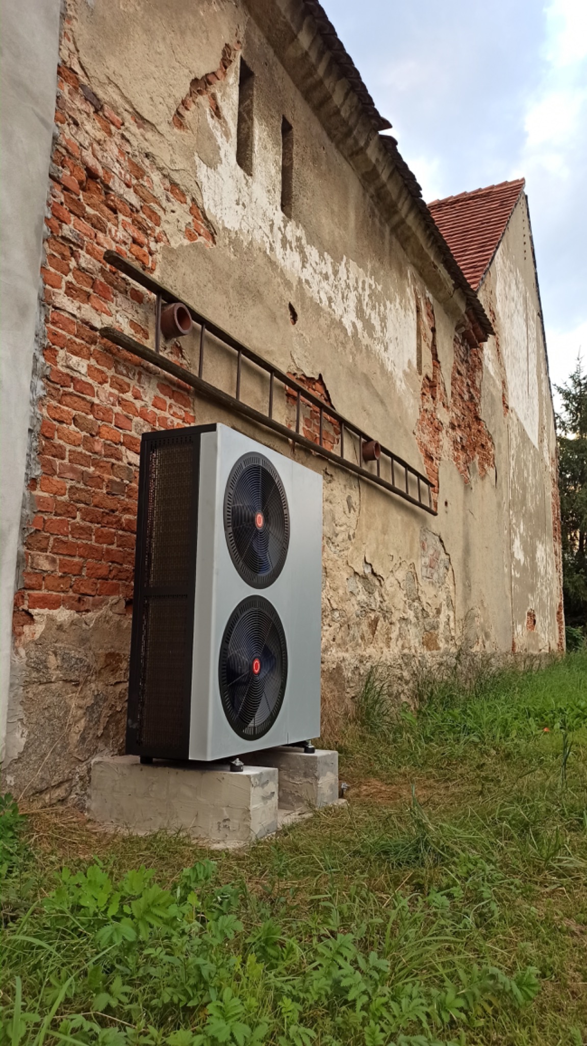 Pompa ciepła powietrze-woda zamontowana na betonowym fundamencie przy starej, ceglanej ścianie budynku z widoczną drabiną.
