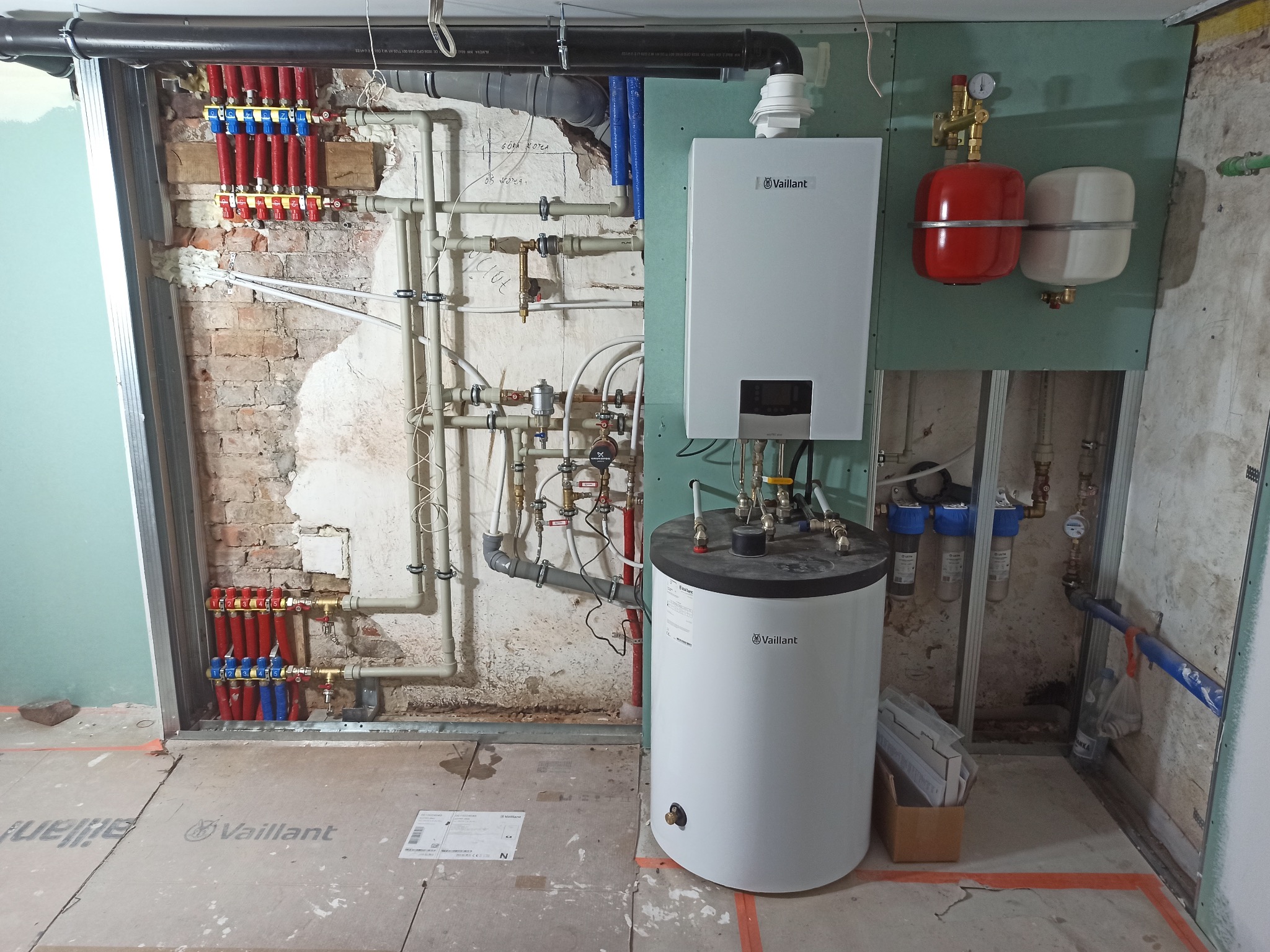 Nowa instalacja grzewcza w trakcie montażu: rozdzielacz podłogówki z czerwono-niebieskimi rurami, wiszący piec gazowy Vaillant, zbiornik buforowy, filtry wody i rury w surowym wnętrzu z fragmentami...