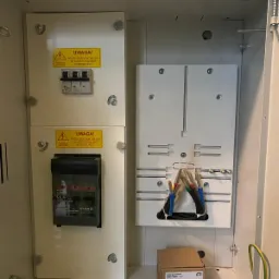 Otwarta skrzynka rozdzielcza z widocznymi bezpiecznikami, przewodami z tulejkami oraz naklejkami ostrzegawczymi.