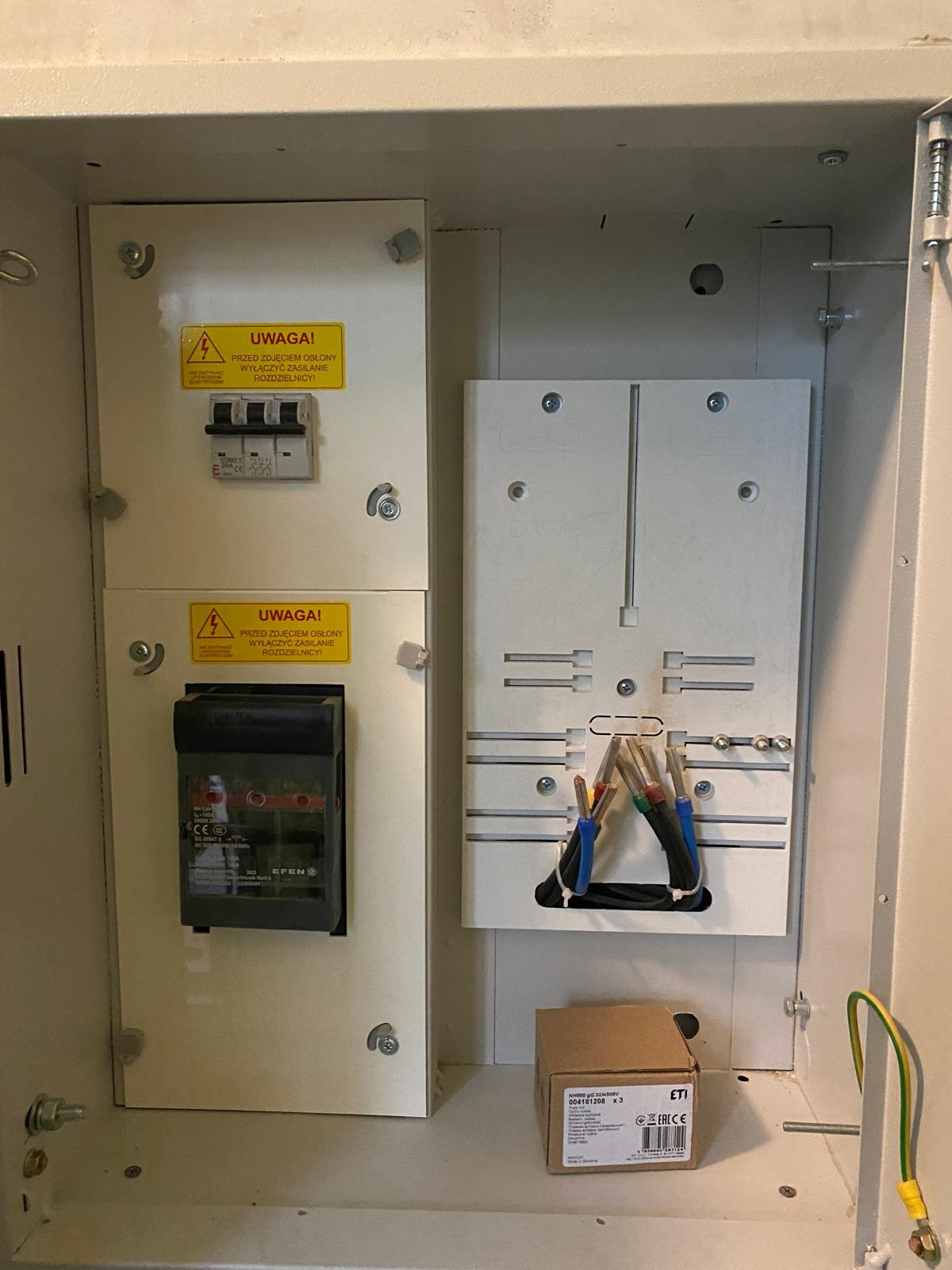 Otwarta skrzynka rozdzielcza z widocznymi bezpiecznikami, przewodami z tulejkami oraz naklejkami ostrzegawczymi.