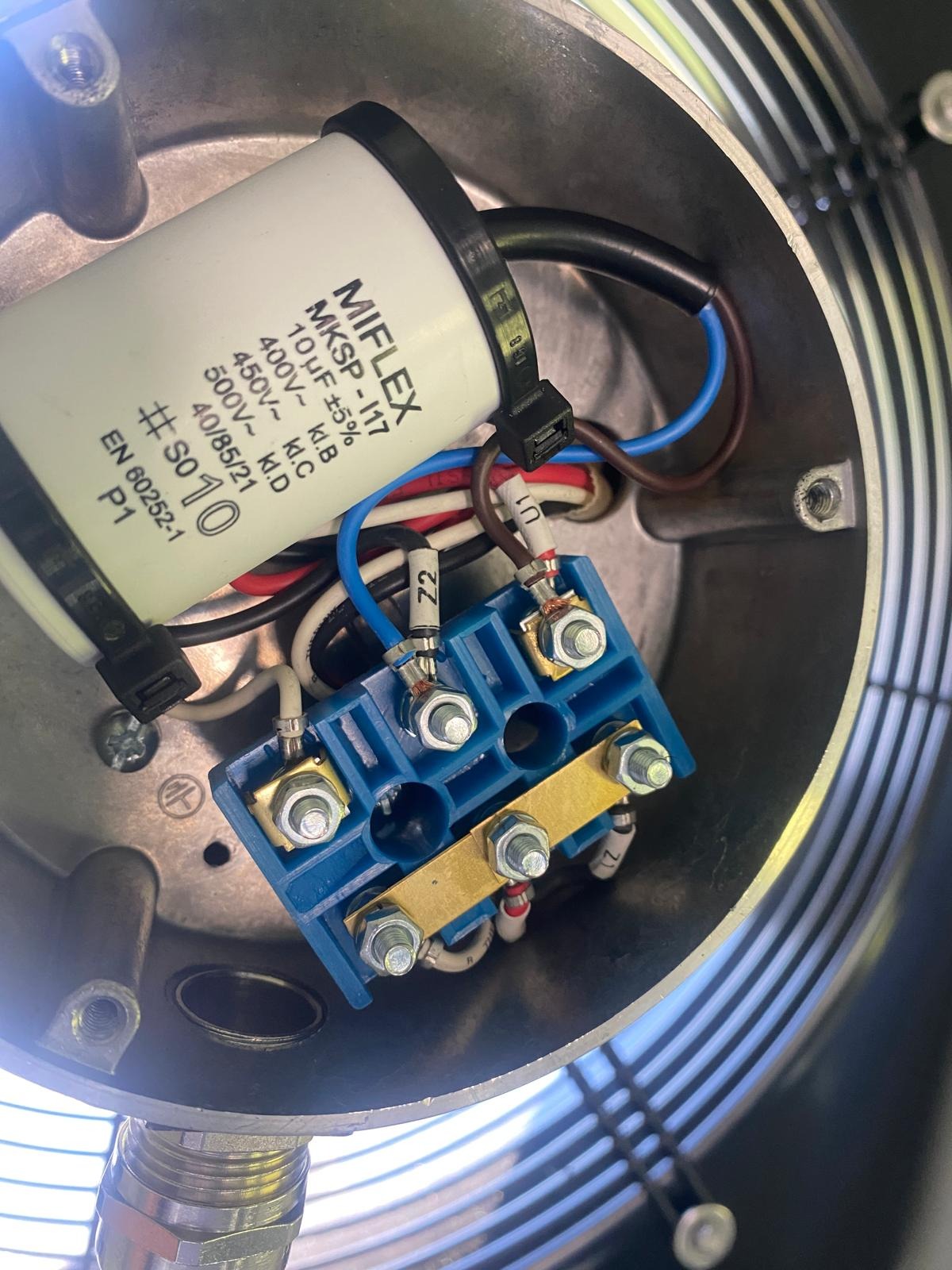 Wnętrze puszki elektrycznej z kondensatorem MIFLEX MKSP-I17, niebieską kostką przyłączeniową z mosiężnymi elementami oraz podłączonymi przewodami o różnych kolorach, widoczne śruby i nakrętki...