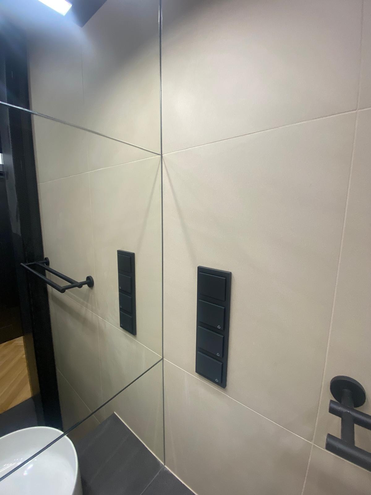 Nowoczesna łazienka z beżowymi płytkami, czarnymi włącznikami światła i relingiem na ręczniki, widoczna w lustrzanym odbiciu.