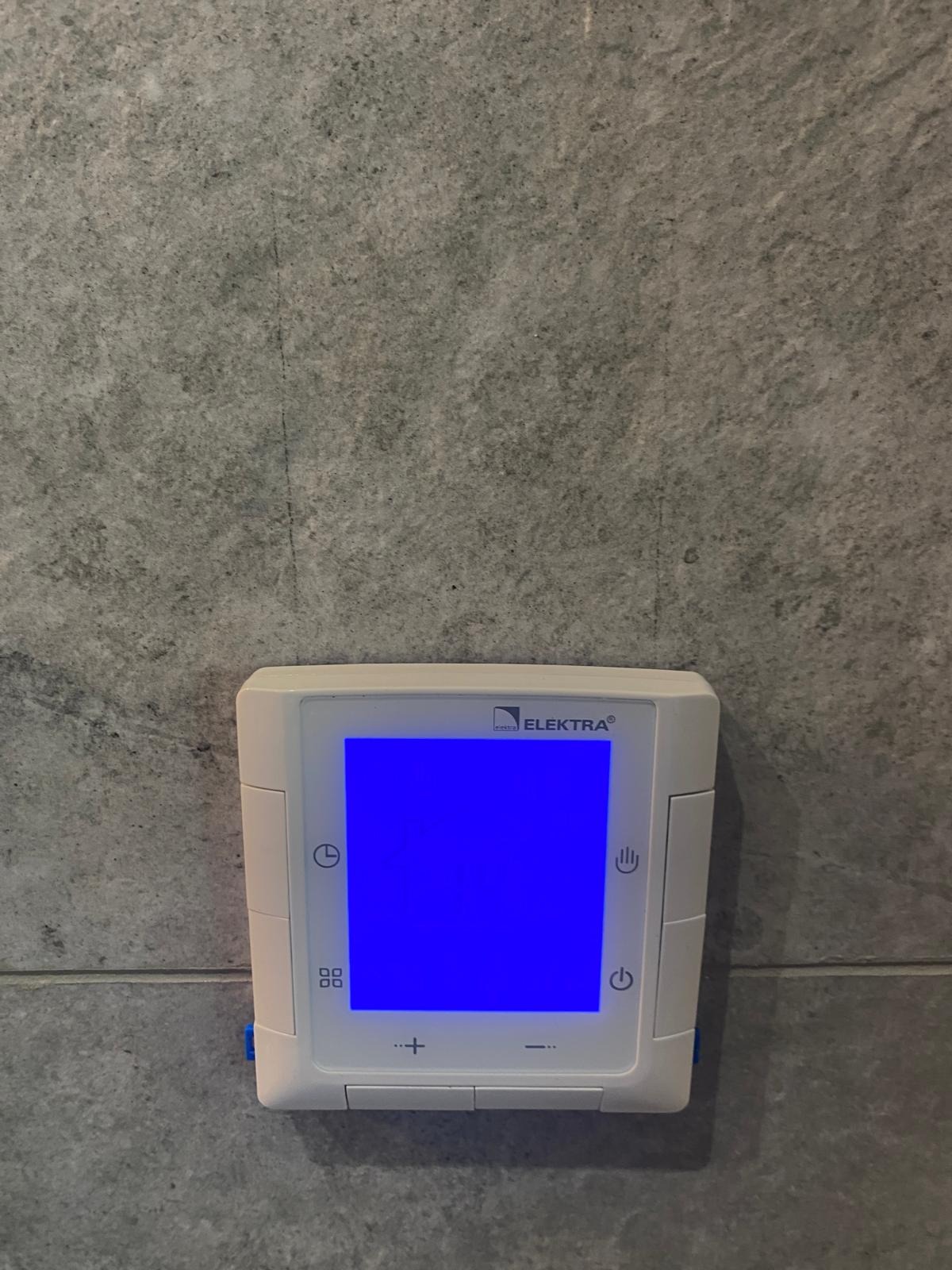 Biały, kwadratowy termostat Elektra zamontowany na szarej ścianie z efektem betonu, z niebieskim ekranem.