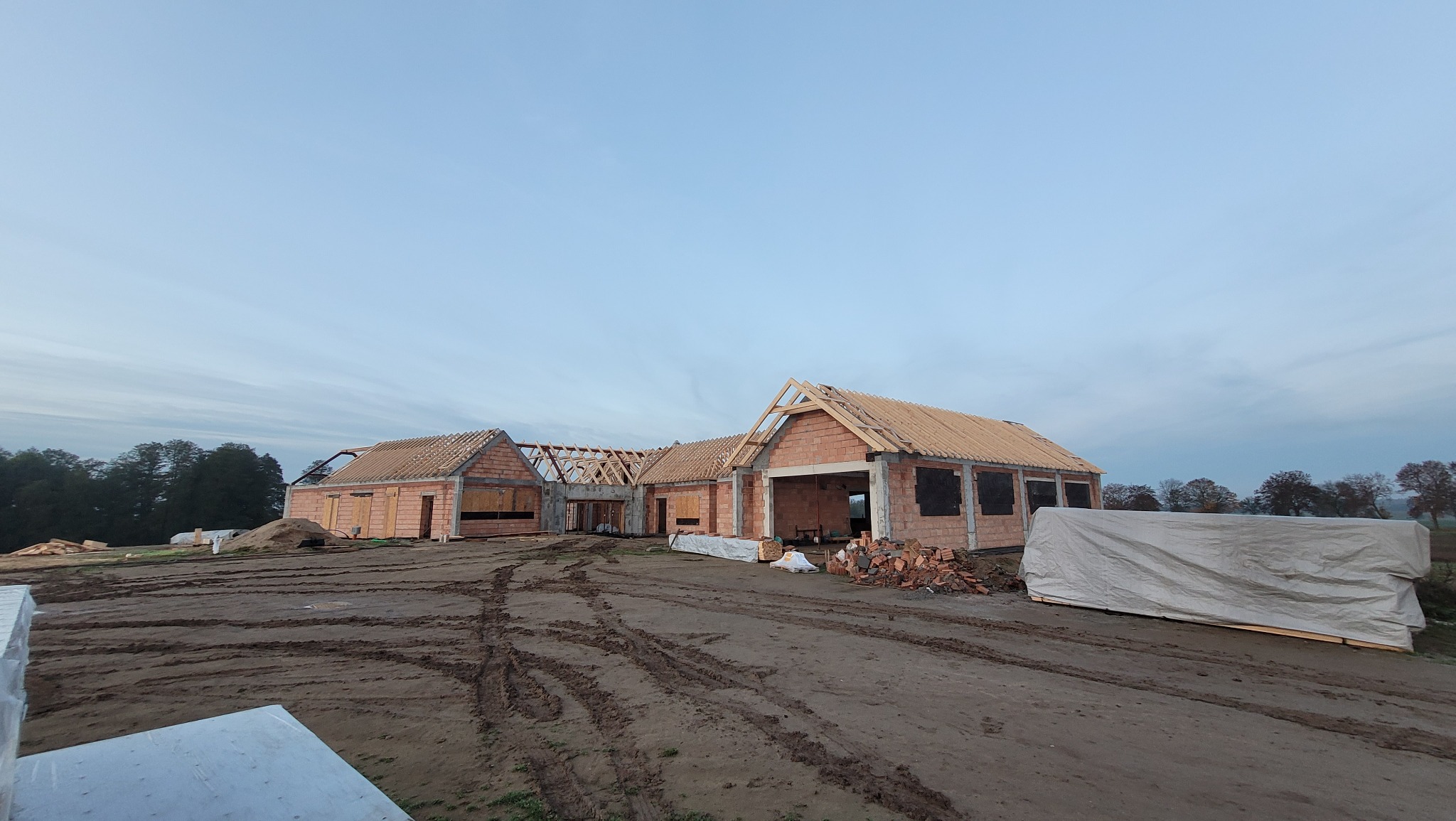 Wznoszenie parterowego domu z czerwonej cegły; widoczne ślady opon na ziemi, drewniana konstrukcja dachu w trakcie montażu.