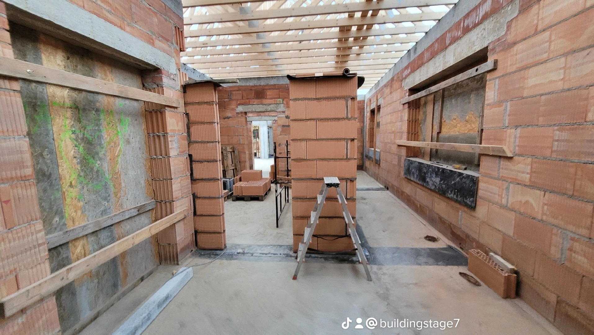 Wnętrze w trakcie budowy z czerwonej cegły, widoczne drewniane belki stropowe, fragmenty ścian z oknami tymczasowo zabezpieczonymi płytami, drabina aluminiowa w centralnej części kadru.