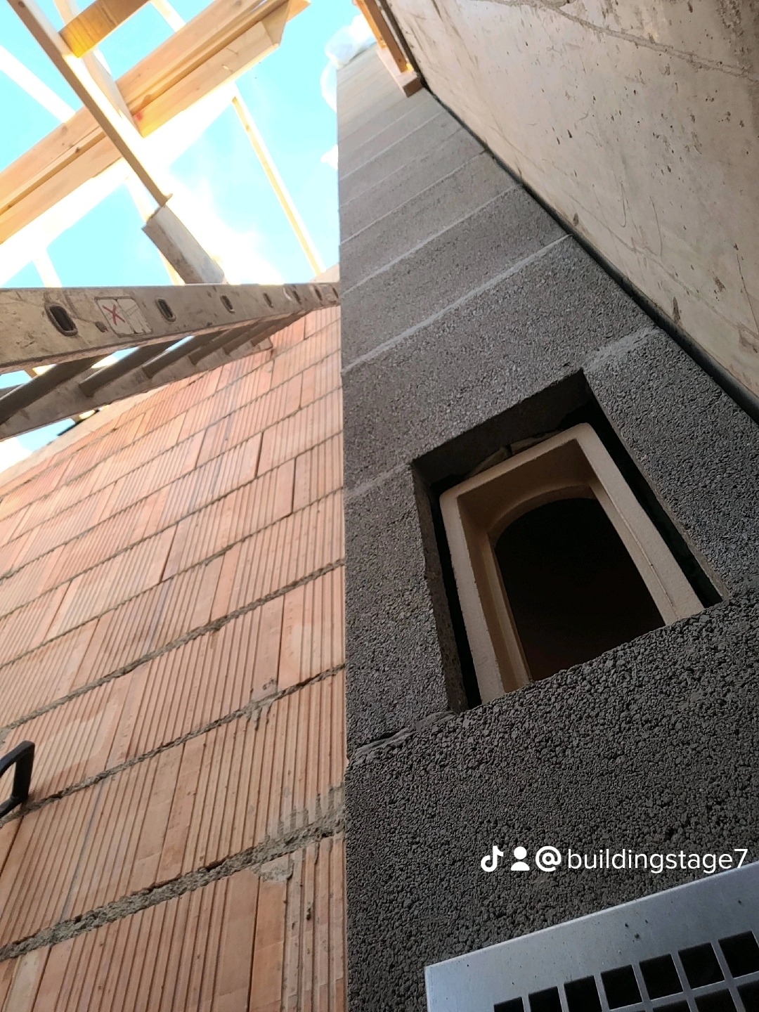 Wnętrze budynku w trakcie budowy: widoczny fragment ściany z czerwonej cegły, szara ściana z otworem okiennym i fragment drewnianej konstrukcji dachu z drabiną na tle błękitnego nieba.