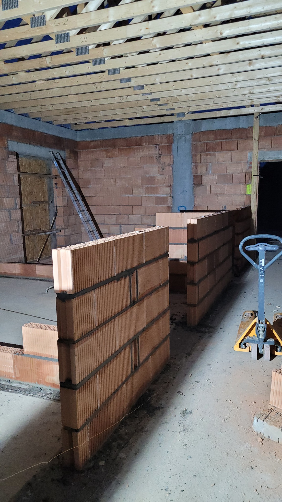 Wznoszenie ścian działowych z czerwonej cegły ceramicznej w surowym stanie, widoczne drewniane belki stropowe, paleciak oraz drabina oparta o ścianę.