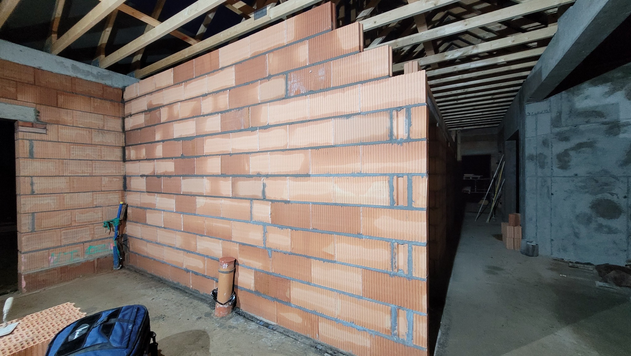 Wnętrze w trakcie budowy: ściana z czerwonej cegły kratówki na poddaszu, widoczne drewniane krokwie dachu, fragment instalacji kanalizacyjnej przy ścianie, torba narzędziowa na podłodze.