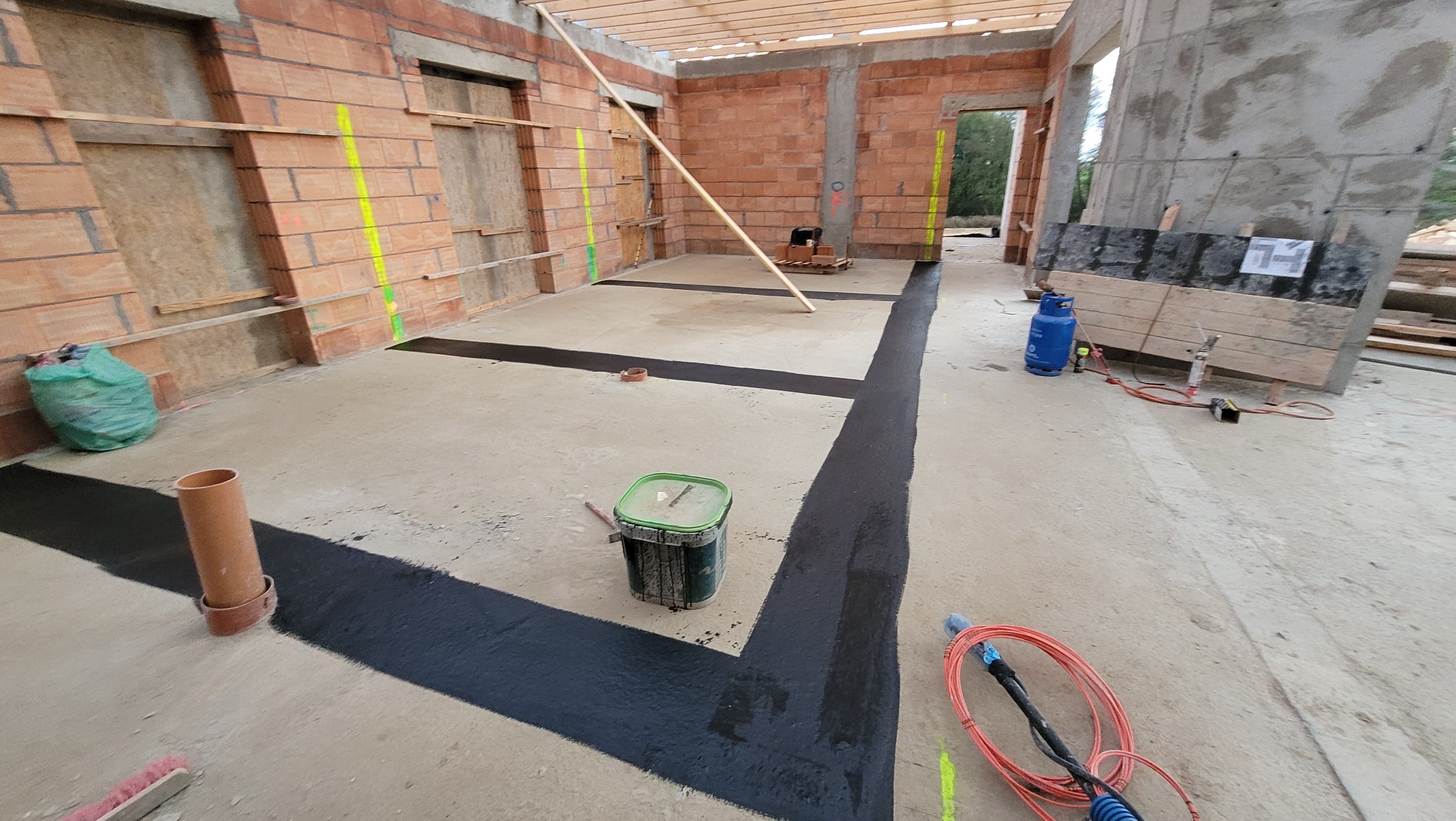 Fundamenty w trakcie izolacji poziomej, widoczne ściany z cegły, narzędzia i materiały budowlane, w tym butla gazowa i pędzle.