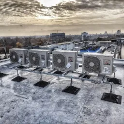 Rząd pięciu zewnętrznych jednostek klimatyzacyjnych Mitsubishi Inverter zamontowanych na metalowej konstrukcji na dachu budynku, z widokiem na panoramę miasta pod pochmurnym niebem.