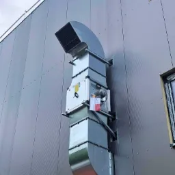 Zewnętrzny kanał wentylacyjny z blachy ocynkowanej, przymocowany do ściany budynku przemysłowego z widocznym silnikiem i dokumentacją techniczną.