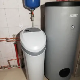 Biały zmiękczacz wody Viessmann obok szarego zasobnika ciepłej wody Emet, z widocznymi miedzianymi rurami instalacji hydraulicznej i niebieskim zbiornikiem wyrównawczym.