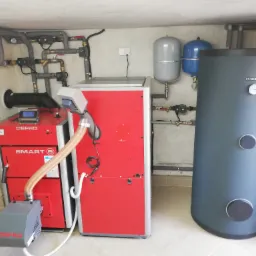 Kotłownia z dwoma czerwonymi kotłami Defro Smart S, szarym zasobnikiem Kospel z zegarem, instalacją rurową pod sufitem i dwoma zbiornikami wyrównawczymi.