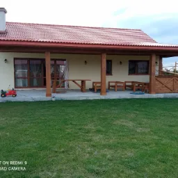 Parterowy dom z czerwonym dachem i drewnianą werandą, na trawniku czerwony traktorek dla dzieci.