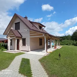 Jasny, parterowy dom z brązowym dachem i roletami zewnętrznymi, widoczny z perspektywy ścieżki z ażurowych płyt betonowych, otoczony trawnikiem i zielenią.