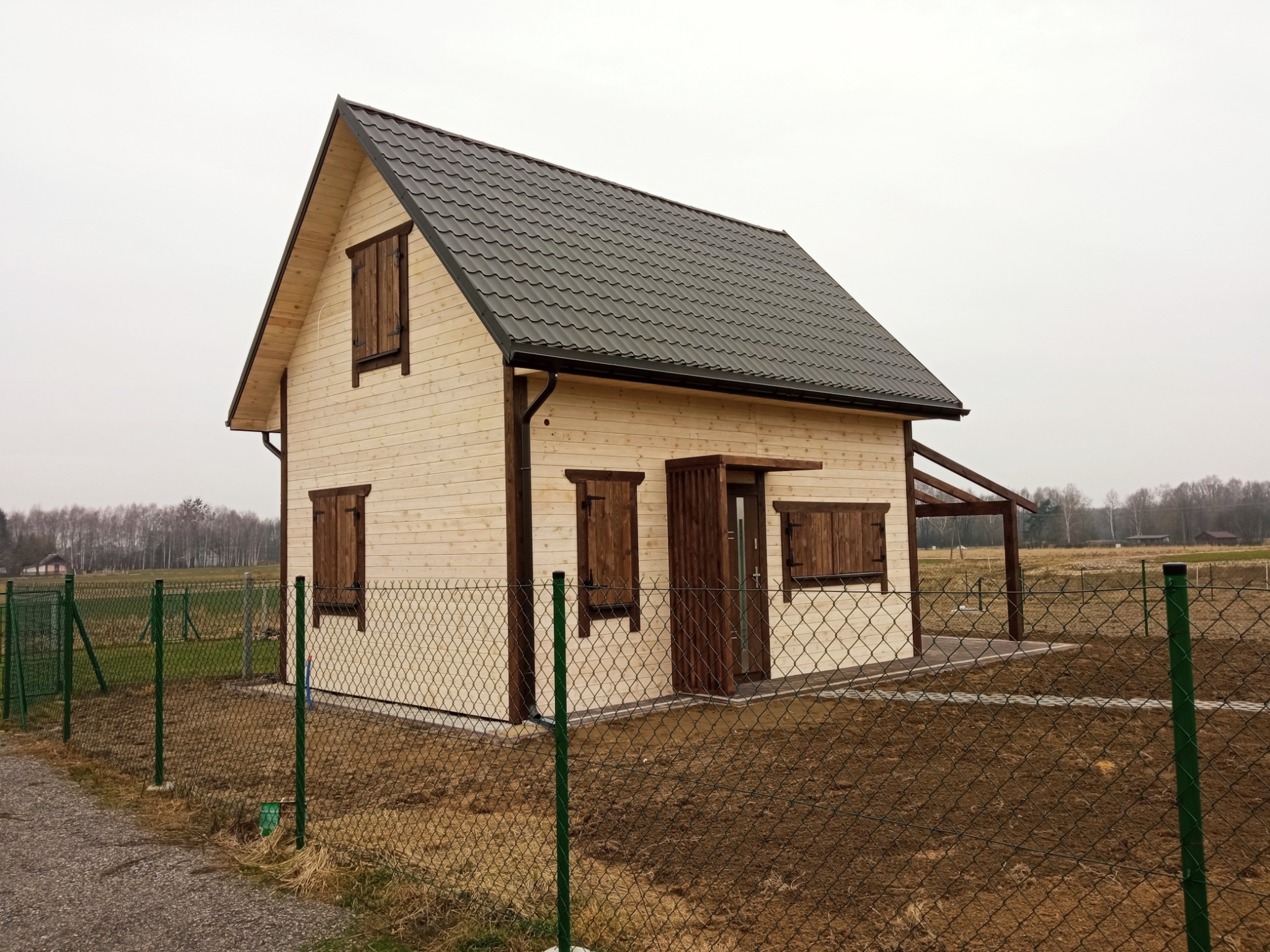 Nowo wybudowany, drewniany dom szkieletowy z ciemnymi okiennicami i grafitowym dachem z blachodachówki, ogrodzony zieloną siatką.