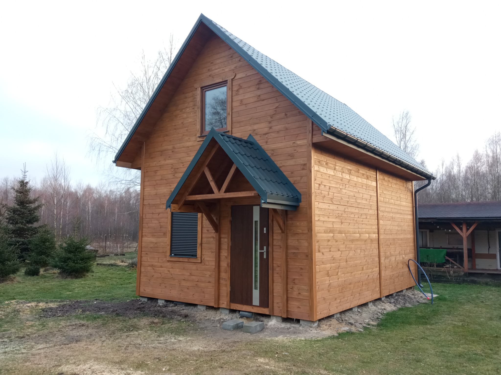 Gotowy, drewniany dom z ciemnym dachem, widok z zewnątrz na tle lasu i pochmurnego nieba. Dom posiada okno, drzwi wejściowe z zadaszeniem i roletę zewnętrzną.