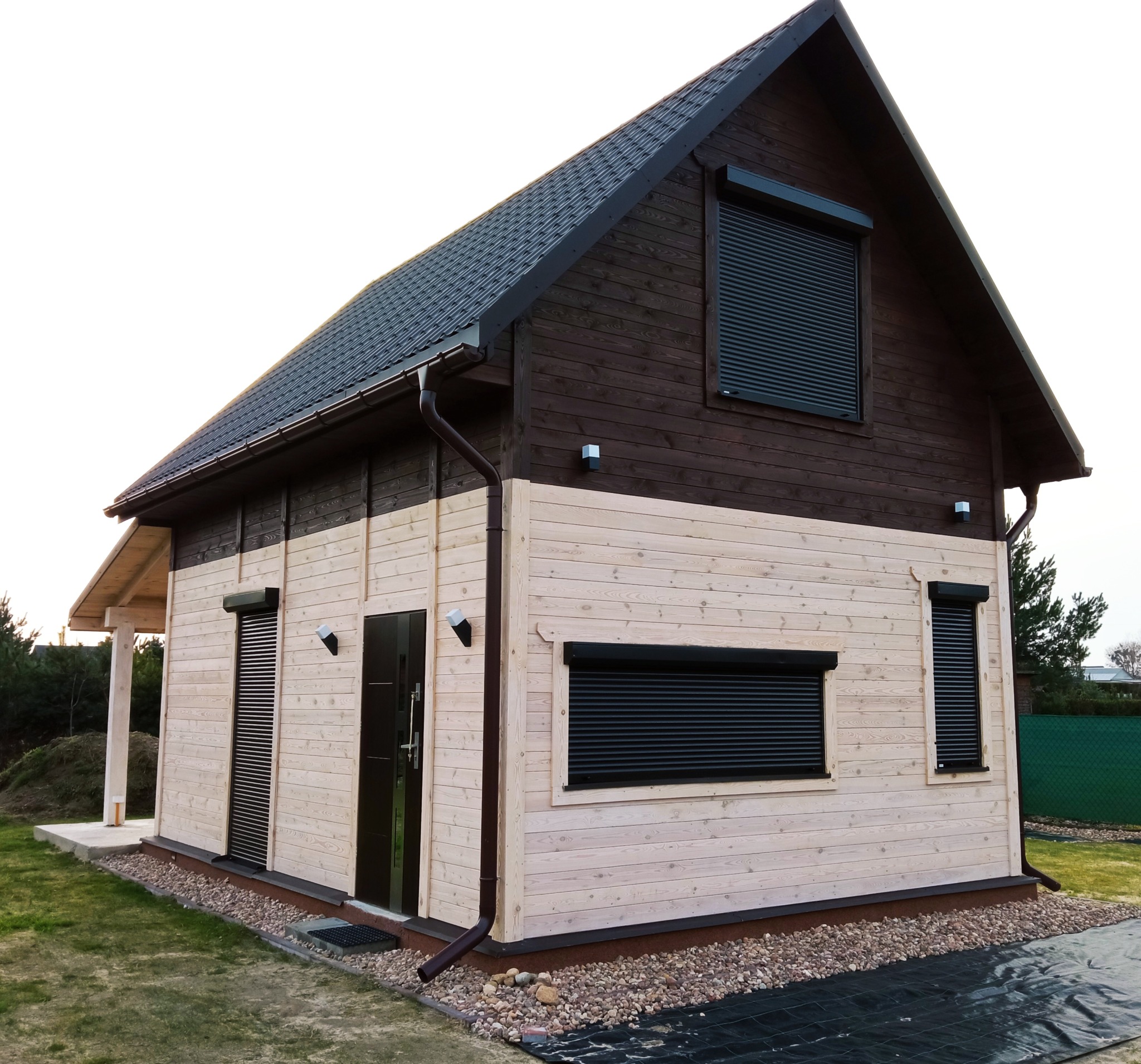 Gotowy dom szkieletowy z ciemnobrązową górną częścią i jasną dolną, z roletami zewnętrznymi, widok z zewnątrz na trawnik i kamienną obwódkę.