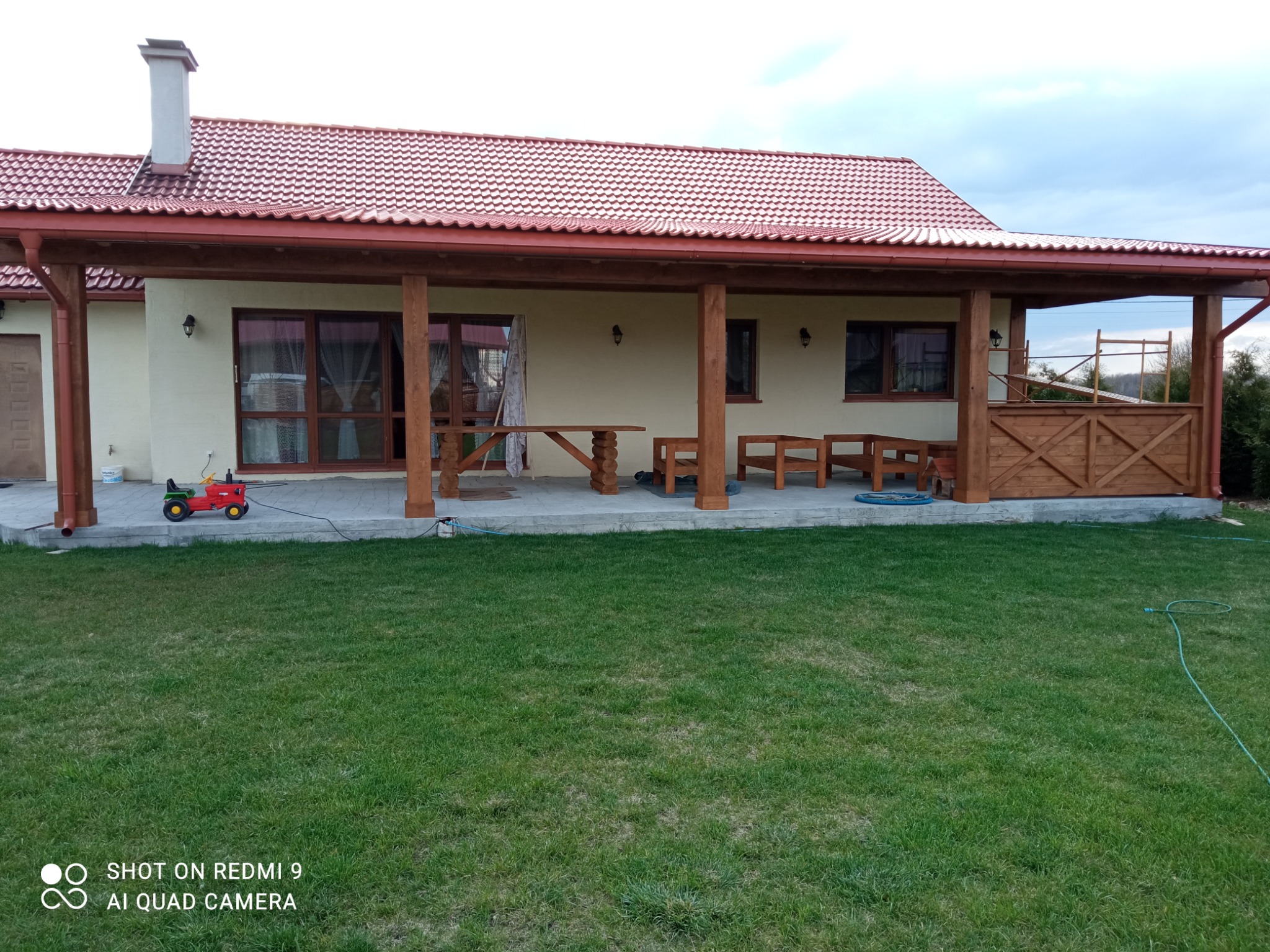 Parterowy dom z czerwonym dachem i drewnianą werandą, na trawniku czerwony traktorek dla dzieci.