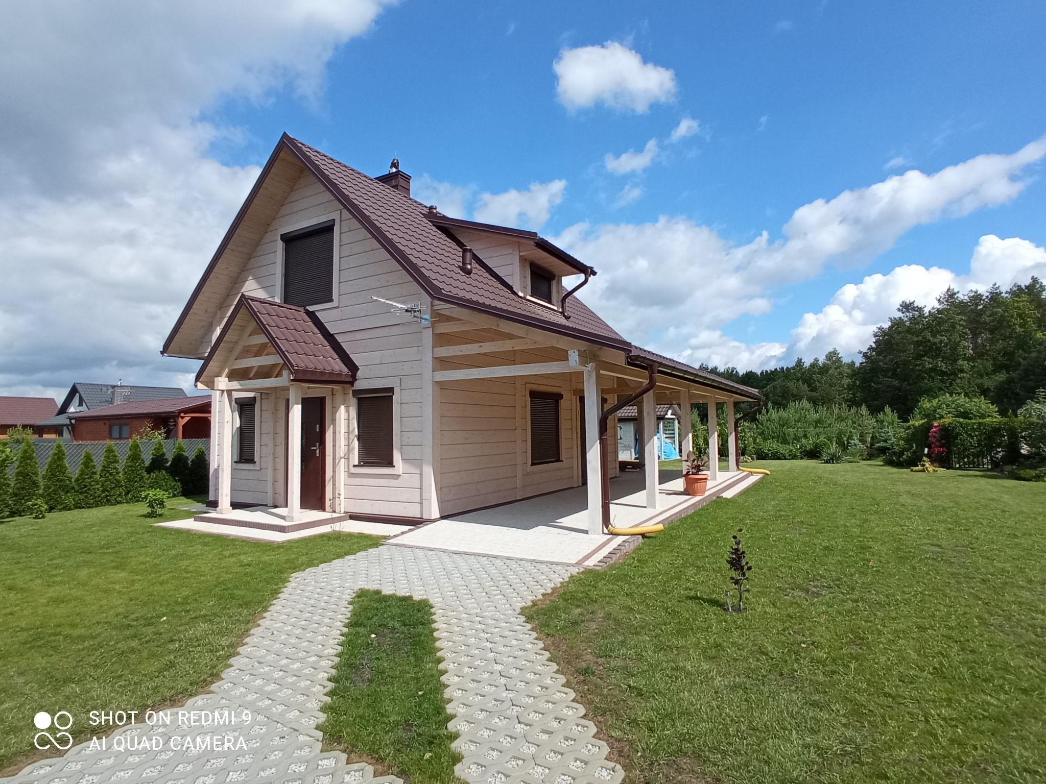 Jasny, parterowy dom z brązowym dachem i roletami zewnętrznymi, widoczny z perspektywy ścieżki z ażurowych płyt betonowych, otoczony trawnikiem i zielenią.