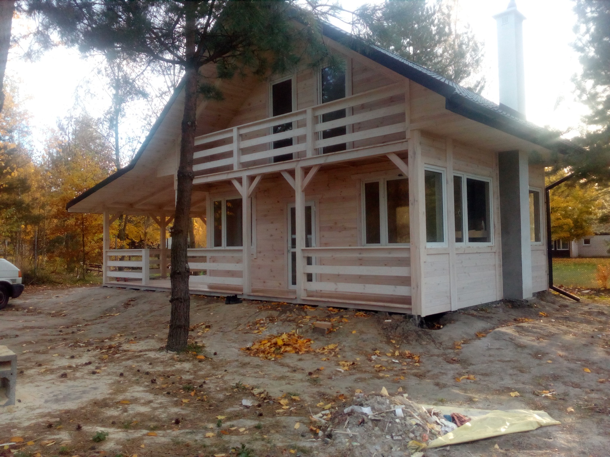 Surowy, drewniany dom z balkonem w stylu skandynawskim, z widocznym kominem i elementami wykończeniowymi, otoczony jesiennym krajobrazem z opadłymi liśćmi.