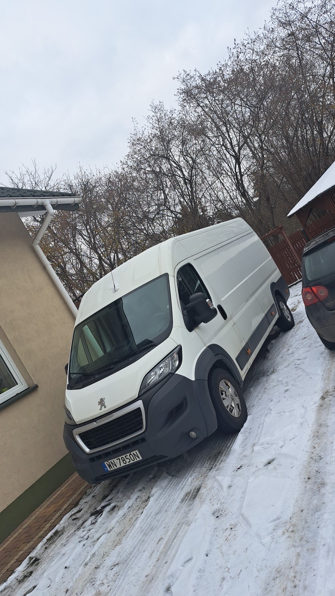 Biały bus Peugeot na śniegu, zaparkowany obok domu z widocznym fragmentem dachu i rynny, tło stanowią drzewa bez liści i fragment zaparkowanego samochodu.