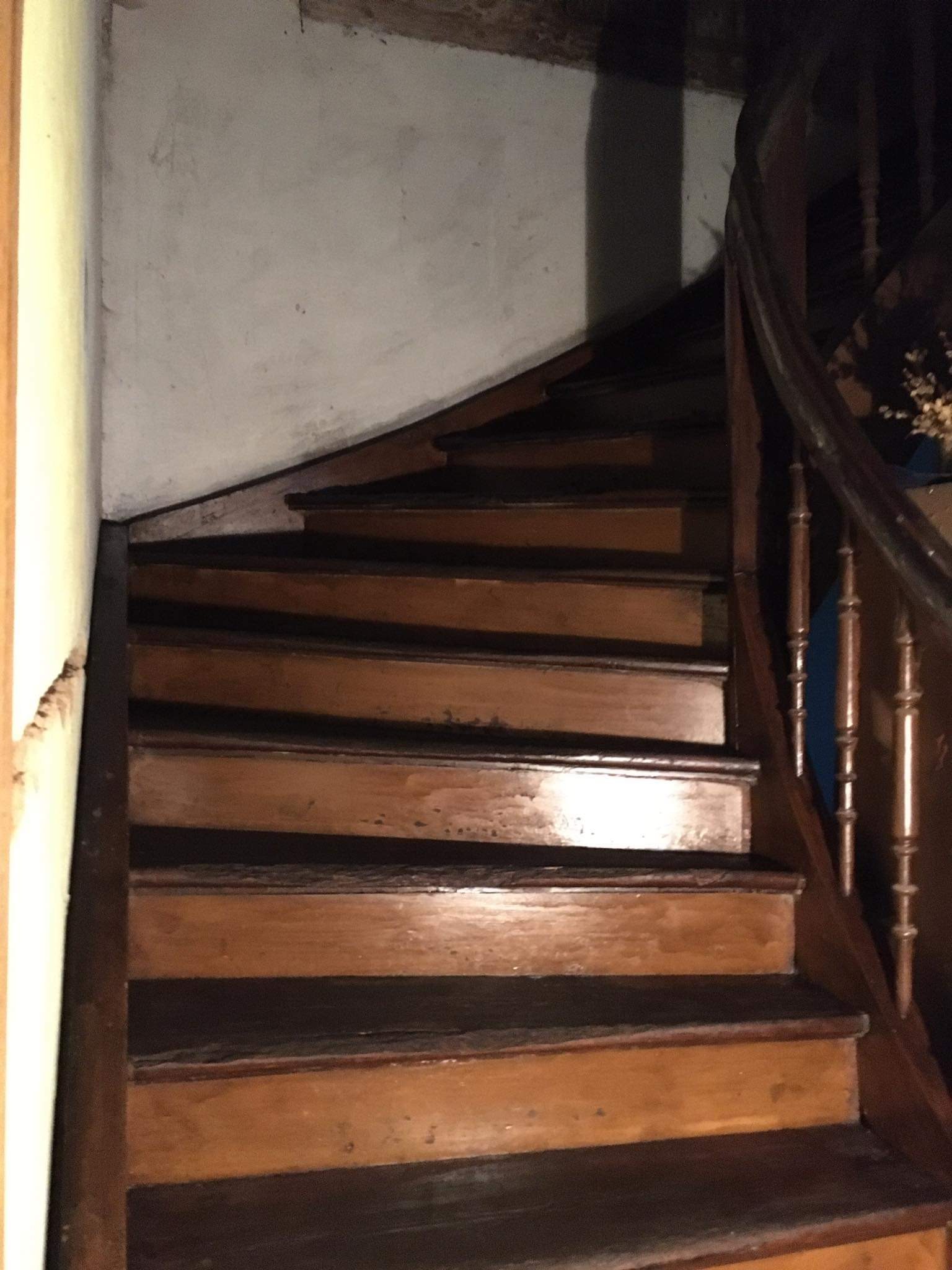 Drewniane, kręcone schody z ozdobną balustradą w słabo oświetlonym wnętrzu, ściany wymagają odświeżenia.
