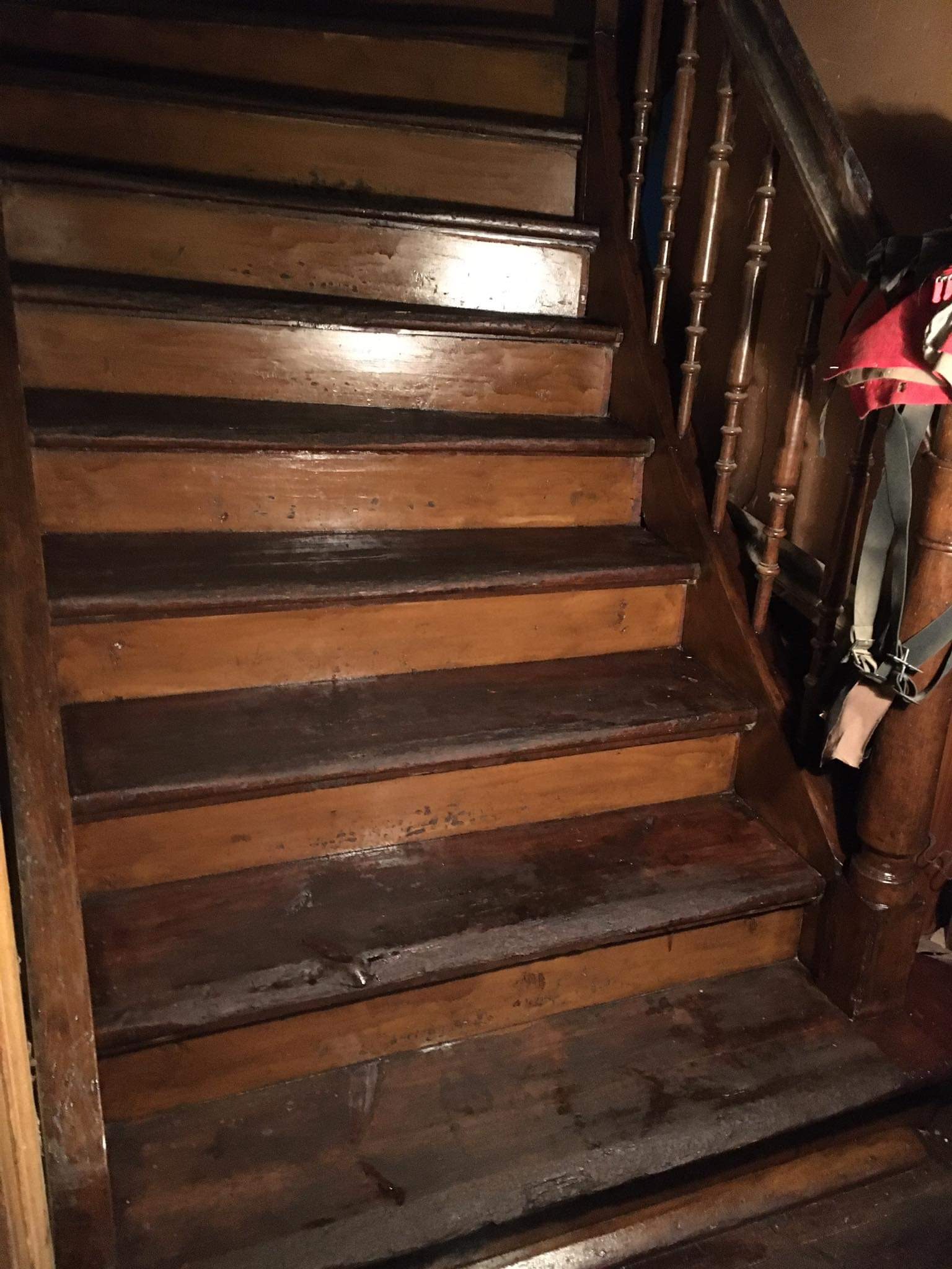 Drewniane schody z ciemnymi stopniami i balustradą, widoczne ślady użytkowania i odświeżania koloru, w tle fragment ściany i czerwona torba.