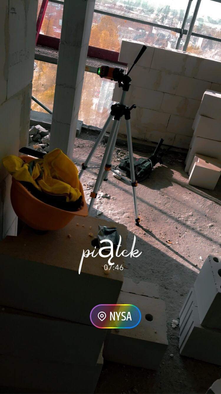 Laserowy niwelator na statywie ustawiony w surowym stanie budynku, obok leży pomarańczowy kask z żółtą bluzą roboczą w środku, w tle widać bloczki z betonu komórkowego.