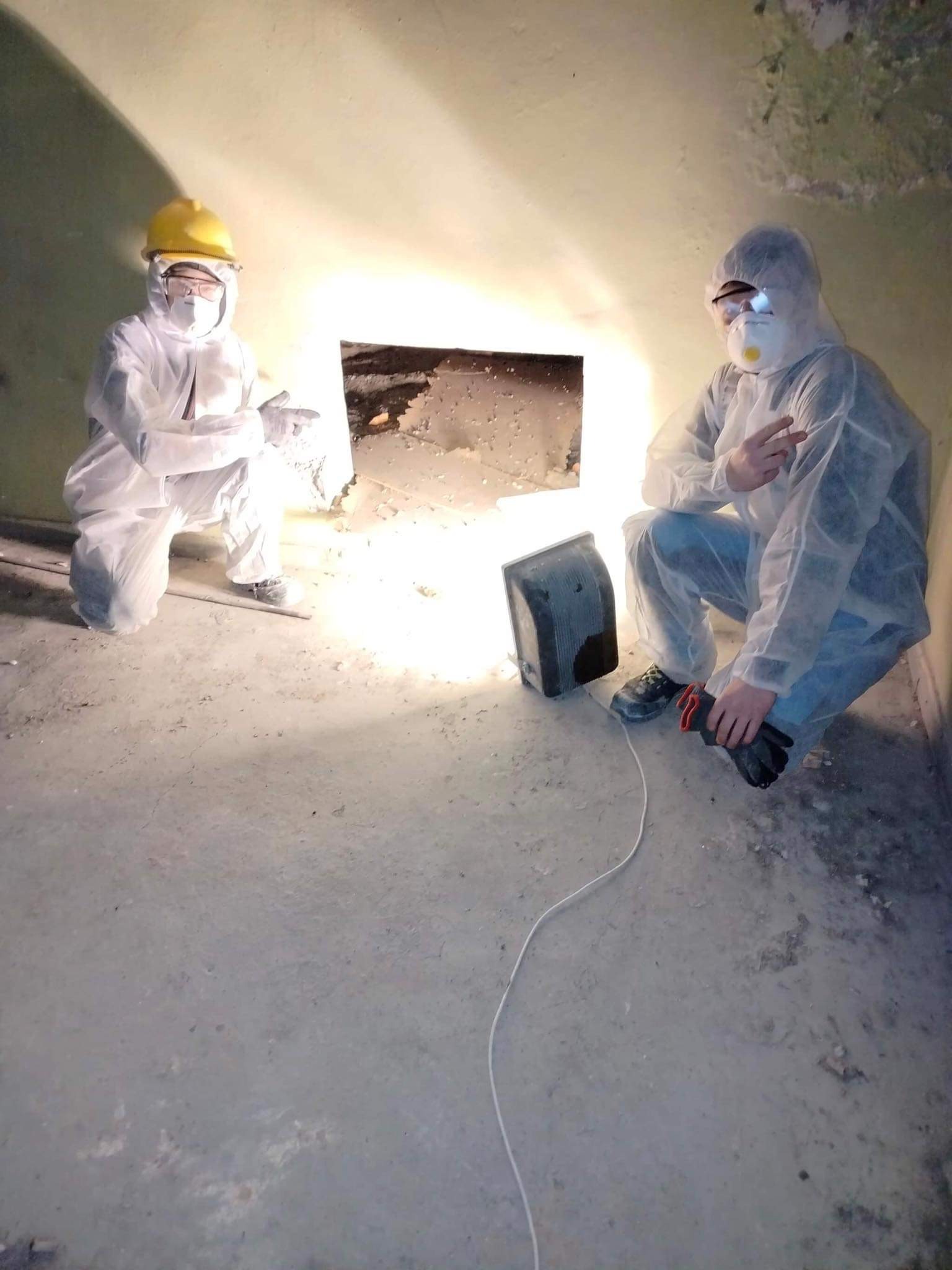 Dwóch pracowników w kombinezonach ochronnych i maskach, jeden w żółtym kasku, oświetla otwór w ścianie podczas prac remontowych w pomieszczeniu.