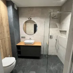 Nowoczesna łazienka z szarymi płytkami imitującymi marmur na podłodze i ścianach, kabina prysznicowa typu walk-in, okrągłe lustro nad umywalką wpuszczoną w drewniany blat, toaleta z białą ceramiką...