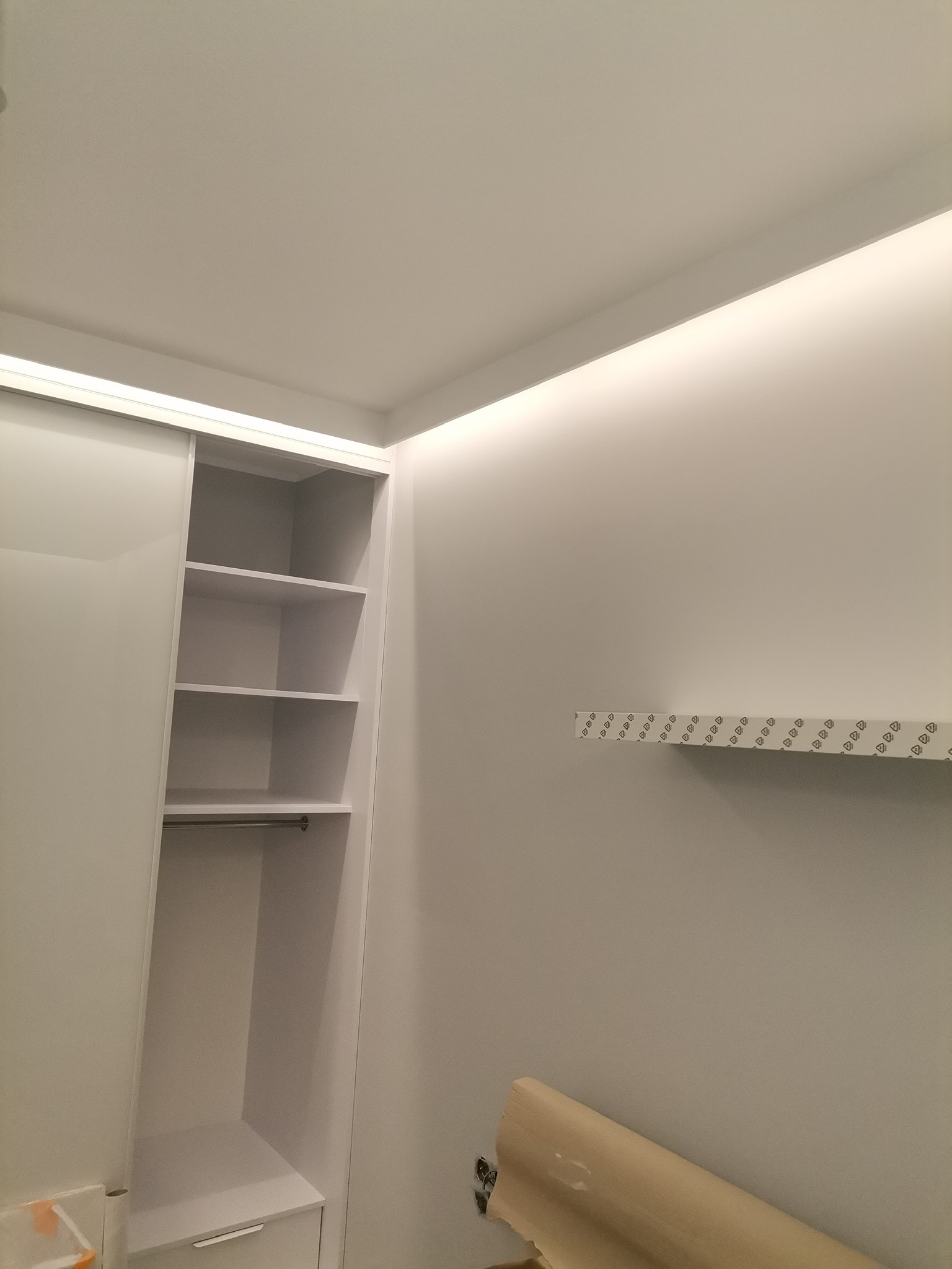 Biała garderoba z przesuwnymi drzwiami i półkami, listwa LED w suficie, zabezpieczona folią podłoga, widoczne ślady po taśmie malarskiej na ścianie