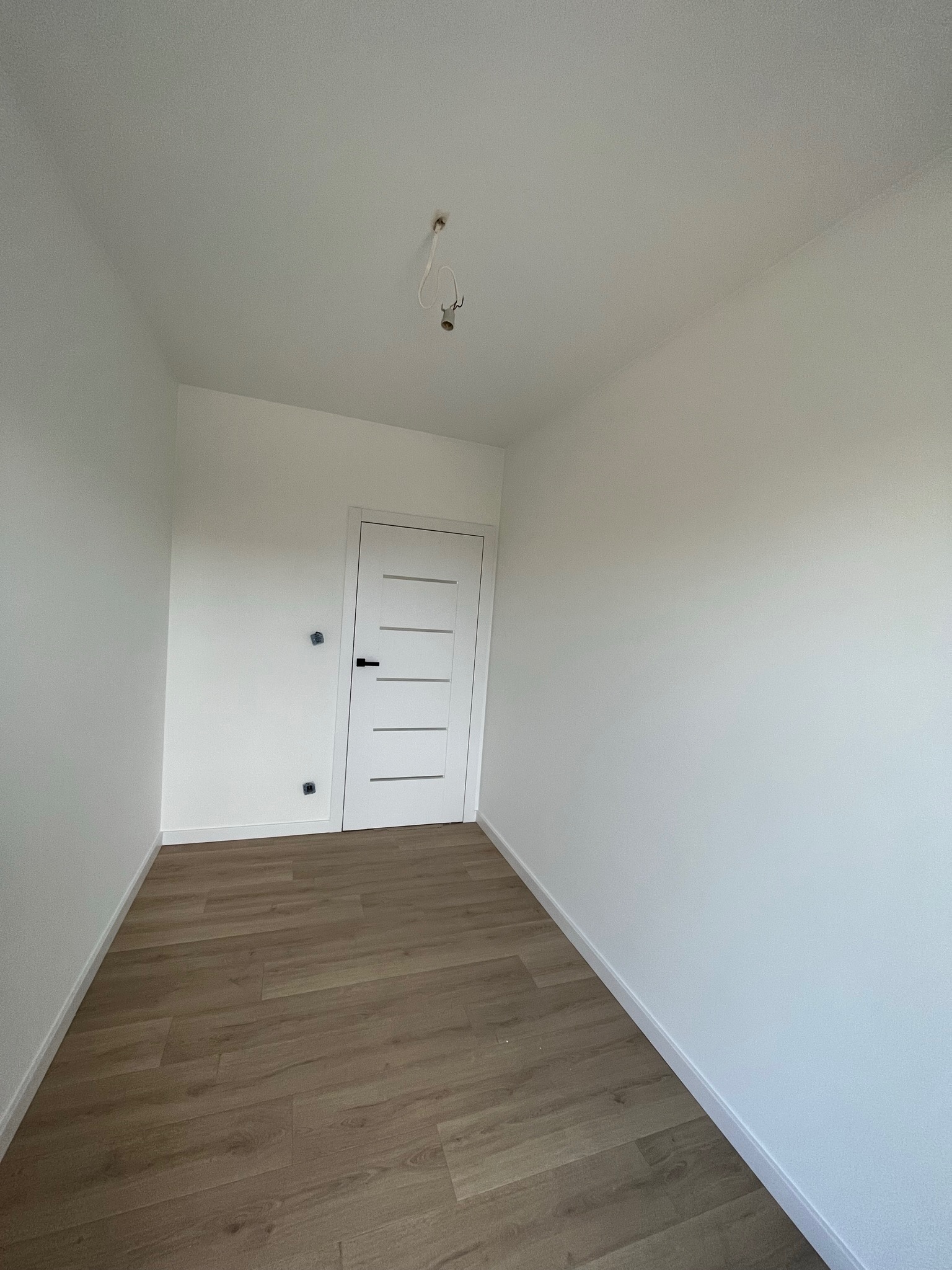 Pusty, świeżo wyremontowany pokój z drewnianą podłogą, białymi ścianami i drzwiami z poziomymi frezami, widoczne wystające kable od oświetlenia sufitowego.
