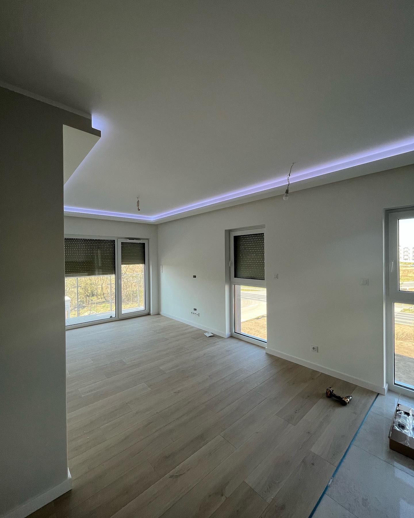 Jasne wnętrze po remoncie z panelami podłogowymi, oświetleniem LED na suficie i dużymi oknami z roletami, widoczne narzędzia i karton na podłodze.