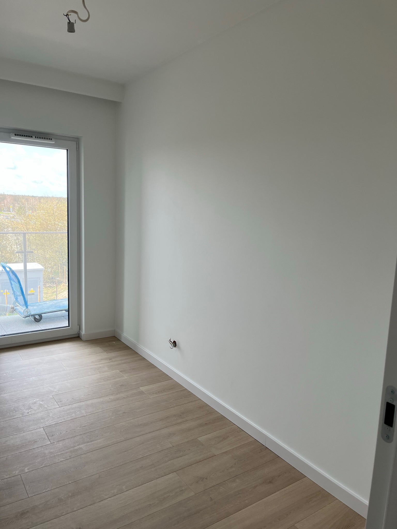 Pomalowane na biało wnętrze pokoju z drewnopodobną podłogą, widoczne gniazdko elektryczne bez osprzętu, białe listwy przypodłogowe i drzwi balkonowe z widokiem na zewnątrz.