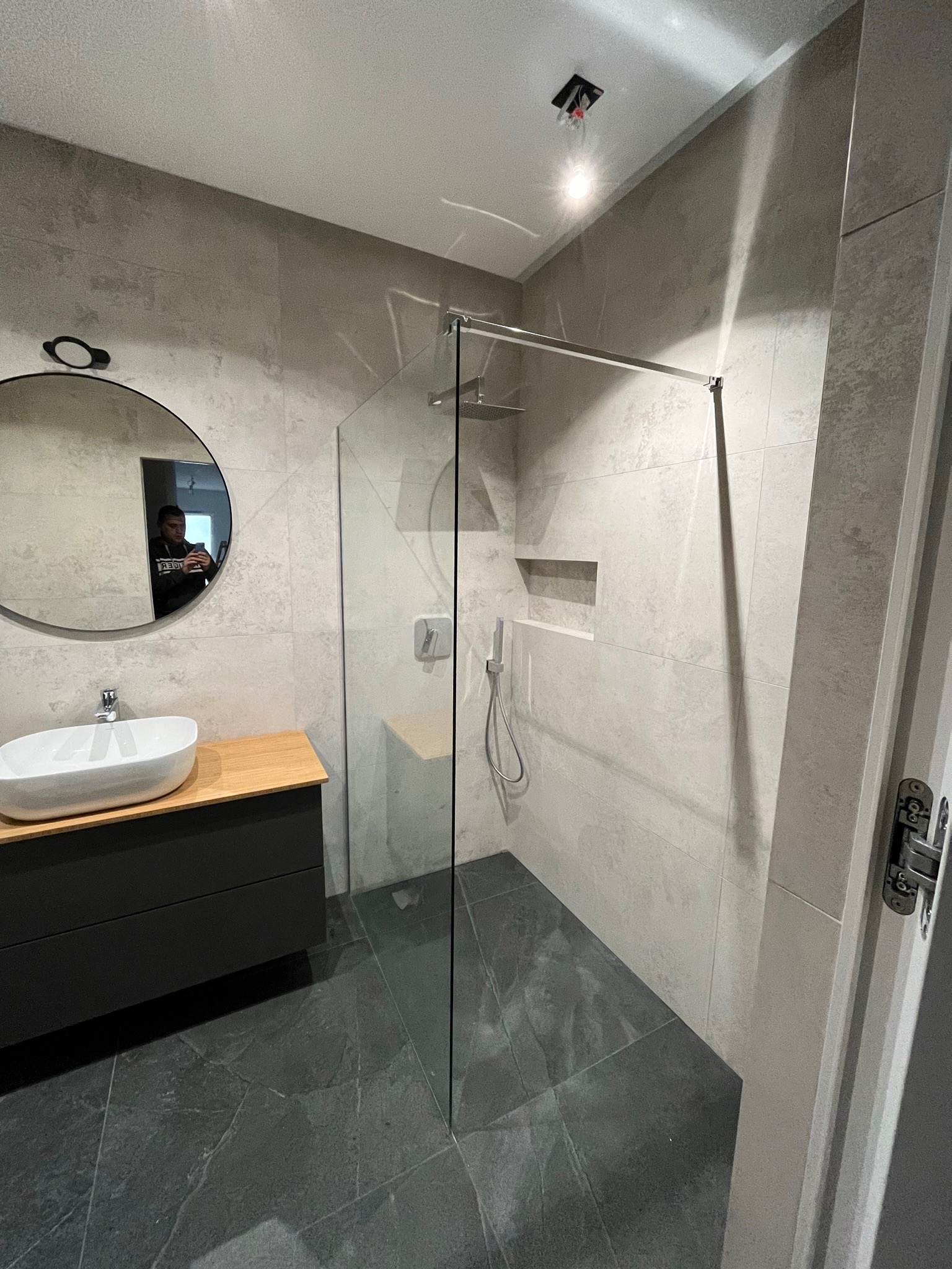 Nowoczesna łazienka z kabiną prysznicową typu walk-in, umywalką nablatową na drewnianym blacie i dużym okrągłym lustrem, wykończona szarymi płytkami w dużym formacie.
