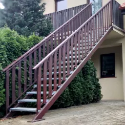 Zewnętrzne schody metalowe z brązowymi barierkami prowadzące na balkon, z widocznymi metalowymi stopniami, otoczone zielonymi krzewami i budynkiem w tle.