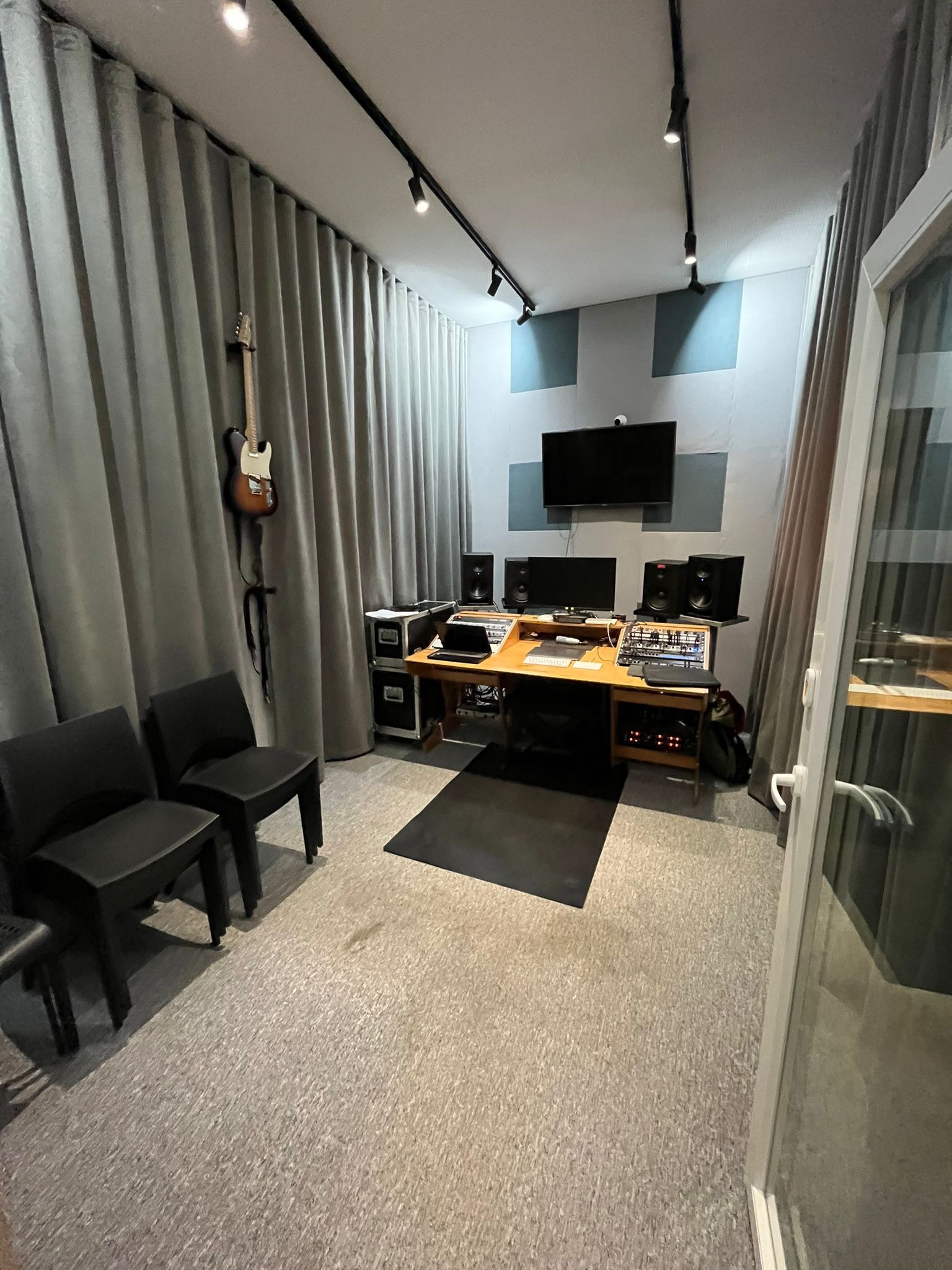Wnętrze studia nagrań z gitarą elektryczną wiszącą na ścianie obok zasłon, biurko z monitorem, głośnikami i mikserem, oświetlenie sufitowe na szynoprzewodach, dwa czarne krzesła.