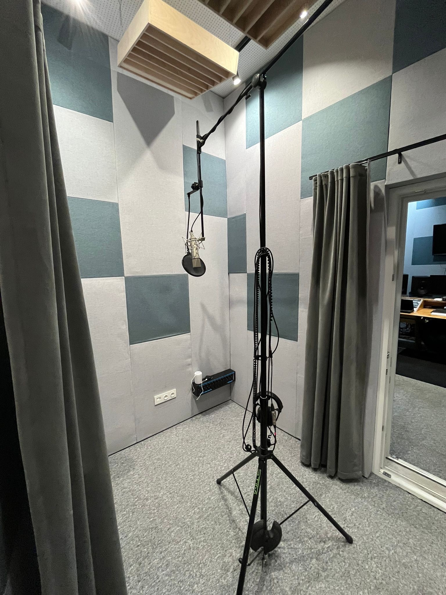 Wnętrze wyciszonego pomieszczenia z mikrofonem studyjnym na statywie, akustycznymi panelami ściennymi w odcieniach szarości i błękitu oraz drewnianym panelem sufitowym.