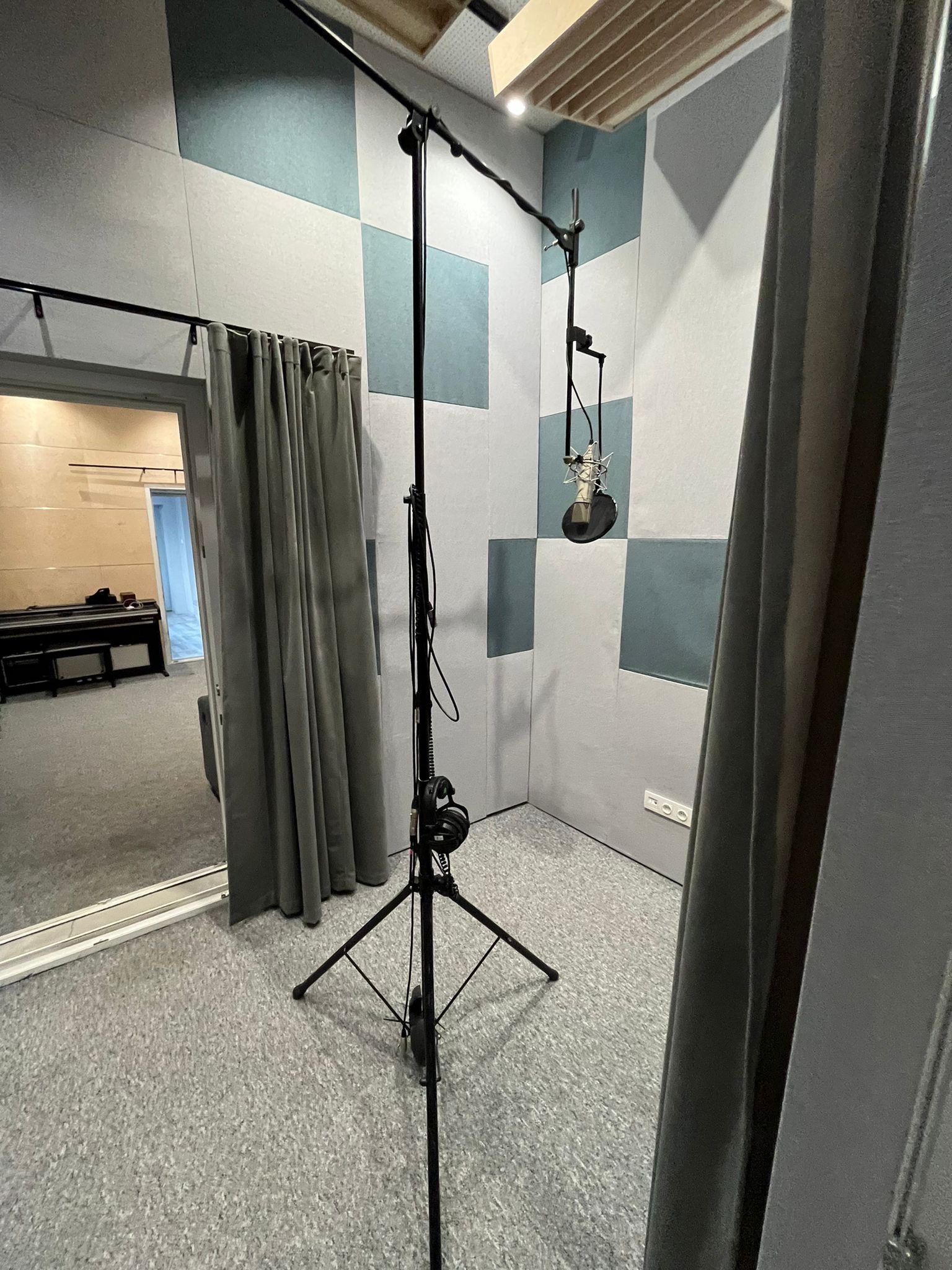 Wnętrze pokoju odsłuchowego z mikrofonem studyjnym zawieszonym na statywie, panele akustyczne na ścianach w szaro-niebieską szachownicę, widoczne wejście do drugiego pomieszczenia z pianinem.