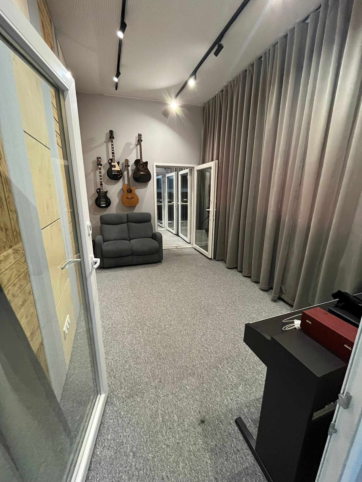 Wnętrze studia nagrań z szarą sofą, kilkoma gitarami wiszącymi na ścianie, oświetleniem punktowym i zasłonami w tle.