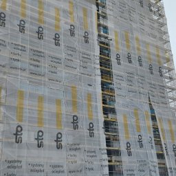 DANI-ELEWACJE - Wysoki budynek w trakcie ocieplania elewacji, osłonięty rusztowaniami i siatką z nadrukiem reklamowym firmy STO, widoczne materiały izolacyjne na placu budowy.