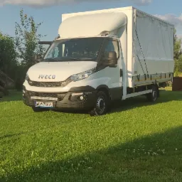 Biały samochód dostawczy Iveco z plandeką, zaparkowany na trawniku w słoneczny dzień.